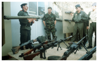 Several military personnel in different uniforms inspect a collection of captured weapons, including various machine guns and a rocket launcher, laid out on a table.